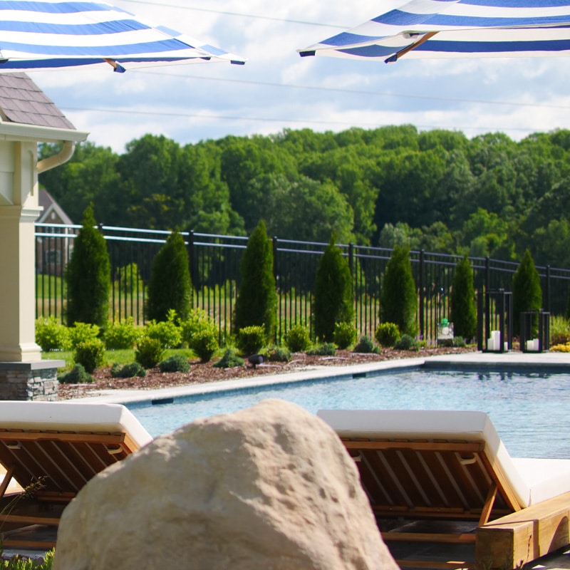 Luxurious poolside scene with lounge chairs, striped umbrellas, and lush greenery under a clear sky.