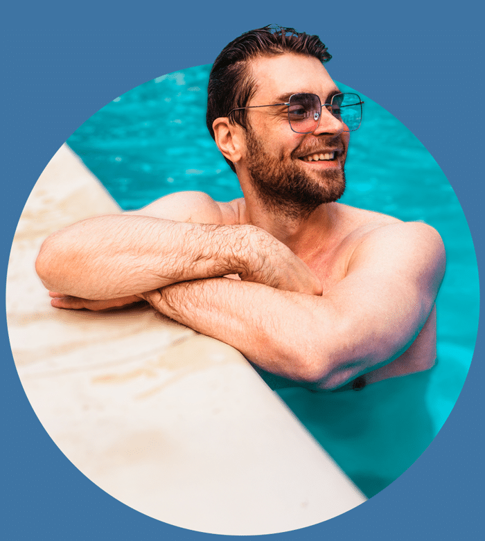 Smiling man wearing sunglasses relaxing in a pool, leaning on poolside with blue water in the background.